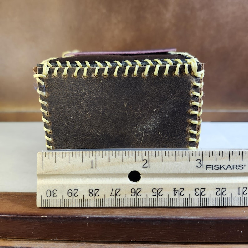 K&ouml;nnte beinhalten: Eine dunkelbraune Lederbox mit sichtbaren cremefarbenen N&auml;hten im Baseball-Stil entlang der Kanten. Ein Fiskars-Lineal liegt davor und zeigt, dass die Box eine Breite von etwa 8,25 cm hat. Die Oberseite ist mit einem dunkelvioletten Lederpaneel versehen.