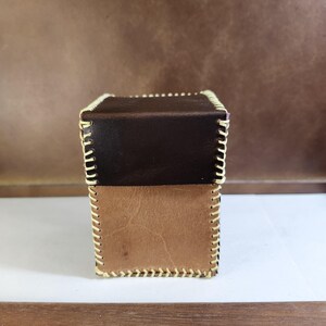 K&ouml;nnte beinhalten: Eine rechteckige Box aus zweifarbigem braunem Leder mit einem dunkelbraunen Oberteil und einem helleren beigen Unterteil. Die Kanten sind mit dicken, cremefarbenen N&auml;hten verbunden, die einen sichtbaren Rand um die gesamte Struktur bilden.