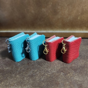 Op de afbeelding: Vier miniatuur leren boeken sleutelhangers. Twee zijn turquoise en twee zijn rood. Elk boek heeft witte pagina's en een metalen sluiting. De turquoise boeken hebben zilveren sluitingen, terwijl de rode boeken gouden sluitingen hebben.