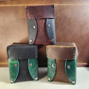 May include: Three handcrafted leather cases in shades of brown, black, and green. Each case features a contrasting flap and is secured with metal snaps. The edges are hand-stitched with a contrasting thread, showcasing the craftsmanship of the design.