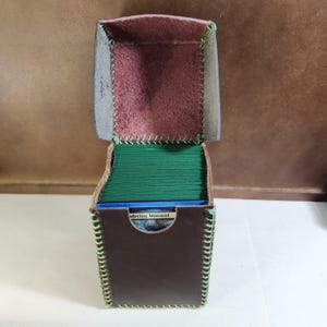 K&ouml;nnte beinhalten: Eine dunkelbraune Leder-Deckbox mit Klappdeckel und hellgr&uuml;nen N&auml;hten an den Kanten. Das Innere ist mit einem strukturierten, rotbraunen Material ausgekleidet. Die Box ist mit einem Stapel gr&uuml;ner Karten gef&uuml;llt, wobei vorne eine Karte mit der Aufschrift Wandering Minstrel zu sehen ist.
