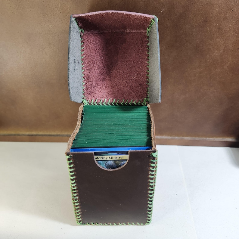 K&ouml;nnte beinhalten: Eine dunkelbraune Leder-Deckbox mit Klappdeckel und hellgr&uuml;nen N&auml;hten an den Kanten. Das Innere ist mit einem strukturierten, rotbraunen Material ausgekleidet. Die Box ist mit einem Stapel gr&uuml;ner Karten gef&uuml;llt, wobei vorne eine Karte mit der Aufschrift Wandering Minstrel zu sehen ist.