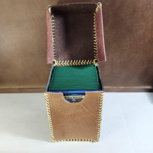 K&ouml;nnte beinhalten: Eine hellbraune Leder-Deckbox mit Klappdeckel und sichtbaren gelben N&auml;hten an den Kanten. Die Box ist mit einem Stapel gr&uuml;n umh&uuml;llter Spielkarten gef&uuml;llt, wobei eine Karte an der Vorderseite den Text Wandering Minstrel aufweist.