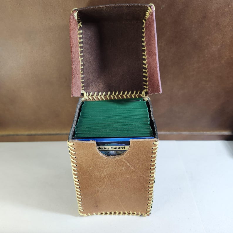 K&ouml;nnte beinhalten: Eine hellbraune Leder-Deckbox mit Klappdeckel und sichtbaren gelben N&auml;hten an den Kanten. Die Box ist mit einem Stapel gr&uuml;n umh&uuml;llter Spielkarten gef&uuml;llt, wobei eine Karte an der Vorderseite den Text Wandering Minstrel aufweist.