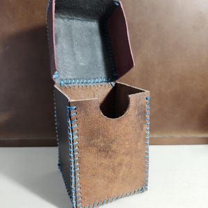 K&ouml;nnte beinhalten: Eine braune Lederbox mit aufklappbarem Deckel und einem halbkreisf&ouml;rmigen Ausschnitt an der Vorderseite. Die Box ist mit sichtbaren, dicken blauen N&auml;hten an allen Kanten versehen, und die Innenseite des Deckels ist in einem dunklen Anthrazit gehalten.