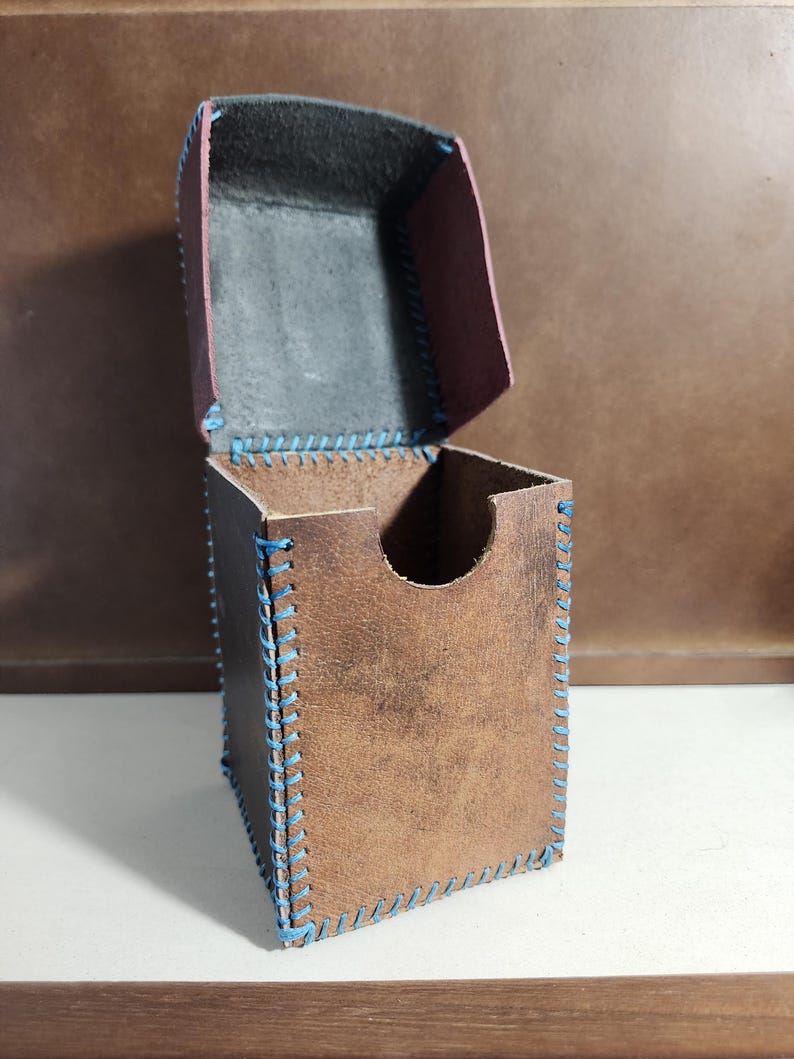 K&ouml;nnte beinhalten: Eine braune Lederbox mit aufklappbarem Deckel und einem halbkreisf&ouml;rmigen Ausschnitt an der Vorderseite. Die Box ist mit sichtbaren, dicken blauen N&auml;hten an allen Kanten versehen, und die Innenseite des Deckels ist in einem dunklen Anthrazit gehalten.