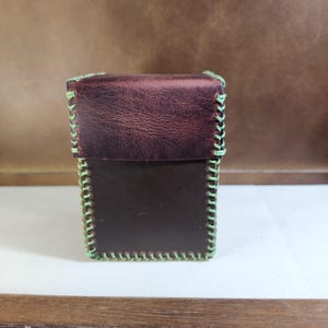 K&ouml;nnte beinhalten: Eine rechteckige Box aus dunkelbraunem Leder mit einem strukturierten Klappdeckel. Die Kanten der Box und des Deckels sind mit sichtbaren, dicken hellgr&uuml;nen N&auml;hten verbunden, die einen markanten Rand um die gesamte Struktur des Beh&auml;lters bilden.