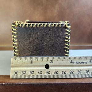 K&ouml;nnte beinhalten: Eine dunkelbraune Lederbox mit sichtbaren gelben N&auml;hten entlang der Kanten. Ein h&ouml;lzernes Fiskars-Lineal ist vor der Box positioniert und zeigt, dass das Objekt eine Breite von etwa 8,9 Zentimetern an der Vorderseite aufweist.