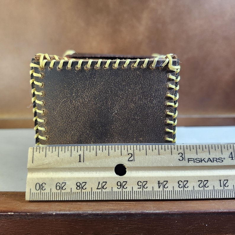 K&ouml;nnte beinhalten: Eine dunkelbraune Lederbox mit sichtbaren gelben N&auml;hten entlang der Kanten. Ein h&ouml;lzernes Fiskars-Lineal ist vor der Box positioniert und zeigt, dass das Objekt eine Breite von etwa 8,9 Zentimetern an der Vorderseite aufweist.