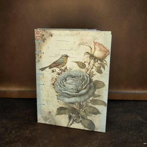 Op de afbeelding: Een klein, handgemaakt dagboek met een vintage bloemenontwerp met een blauwe roos, een roze roos en een kleine vogel. De omslag is gemaakt van papier met een textuur.