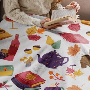 Puede incluir: Una manta blanca con un patr&oacute;n de oto&ntilde;o que presenta ilustraciones de libros, copas de vino, bellotas, peras, teteras y hojas.