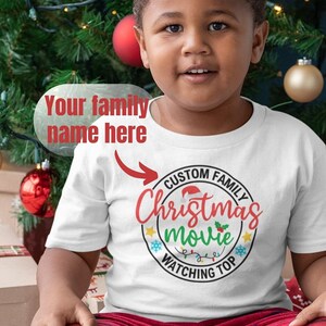May include: A young child wearing a white t-shirt with a red and green Christmas movie watching party design. The shirt says "Custom Family Christmas Movie Watching Top". The child is sitting on a wooden floor with a wrapped gift box in front of them.