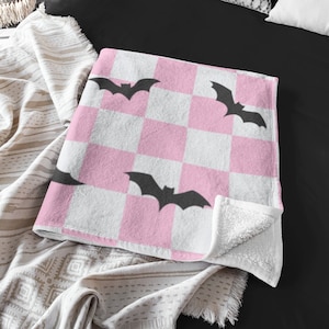 May include: Pink and white checkered throw blanket with black bat silhouettes. The blanket has a soft, plush texture and is perfect for adding a touch of spooky fun to any room.