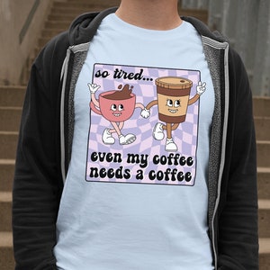 May include: A light blue t-shirt with a cartoon illustration of two coffee cups. The text on the shirt reads "so tired... even my coffee needs a coffee".