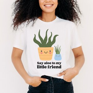 May include: A white t-shirt with a graphic of two potted plants. The larger plant is green and has a yellow pot with a smiling face. The smaller plant is green and has a blue pot with a smiling face. The text "Say aloe to my little friend" is printed below the plants.