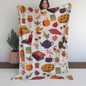Puede incluir: Una manta blanca con un estampado colorido de tem&aacute;tica oto&ntilde;al. El estampado incluye calabazas, hojas, bellotas, tazas de t&eacute;, libros y copas de vino.