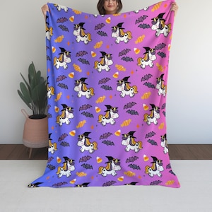 Puede incluir: Una manta de tiro de color morado y azul degradado con un patrón repetido de unicornios blancos con sombreros de bruja y murciélagos. Los unicornios sostienen maíz dulce y los murciélagos vuelan.