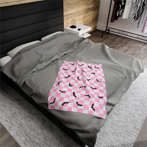 May include: A pink and white checkered throw blanket with black bat silhouettes. The blanket is folded and placed on a gray bed.