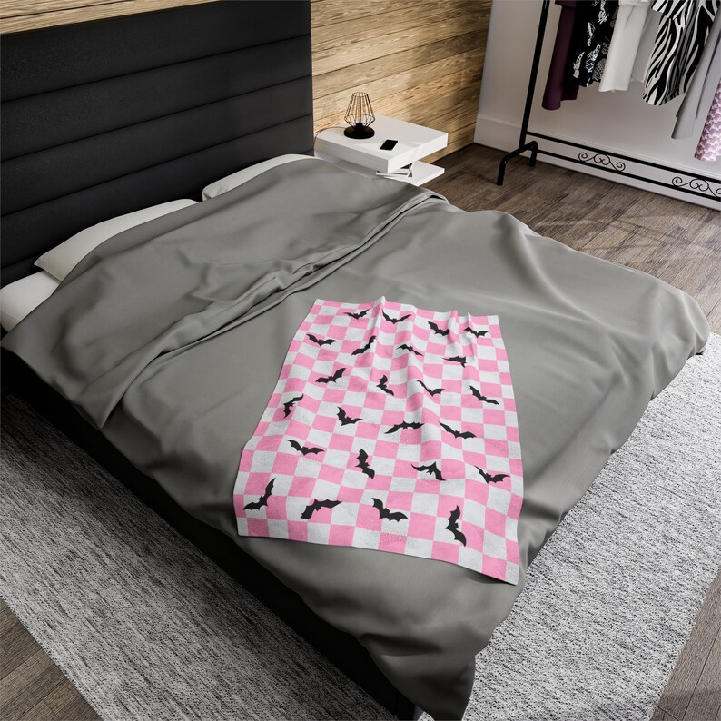 May include: A pink and white checkered throw blanket with black bat silhouettes. The blanket is folded and placed on a gray bed.