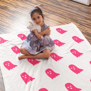 Puede incluir: Una manta blanca con un patr&oacute;n de fantasmas rosas. Una ni&ntilde;a est&aacute; sentada en la manta y sostiene un unicornio de peluche blanco.