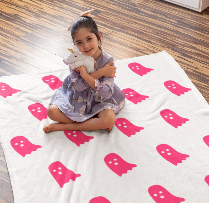 Puede incluir: Una manta blanca con un patr&oacute;n de fantasmas rosas. Una ni&ntilde;a est&aacute; sentada en la manta y sostiene un unicornio de peluche blanco.