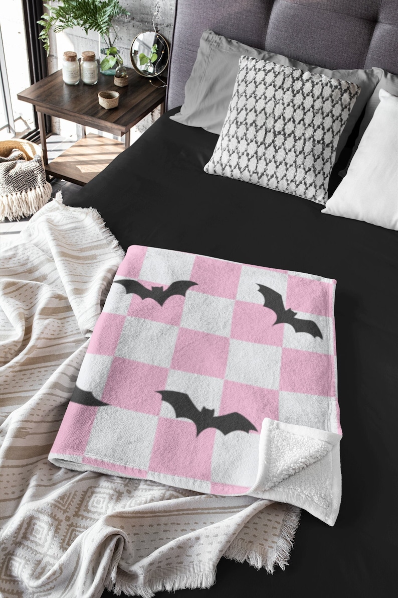 May include: Pink and white checkered throw blanket with black bat silhouettes. The blanket has a soft, plush texture and is perfect for adding a touch of spooky fun to any room.