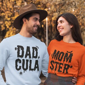May include: A couple wearing matching Halloween themed sweatshirts. The man is wearing a light blue sweatshirt with the text "DAD-CULA" in black dripping letters. The woman is wearing an orange sweatshirt with the text "MOM-STER" in black dripping letters.