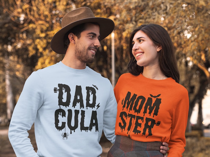 May include: A couple wearing matching Halloween themed sweatshirts. The man is wearing a light blue sweatshirt with the text "DAD-CULA" in black dripping letters. The woman is wearing an orange sweatshirt with the text "MOM-STER" in black dripping letters.
