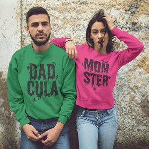 May include: A couple wearing matching green and pink sweatshirts with Halloween-themed text. The green sweatshirt says "Dad-Cula" and the pink sweatshirt says "Mom-ster".