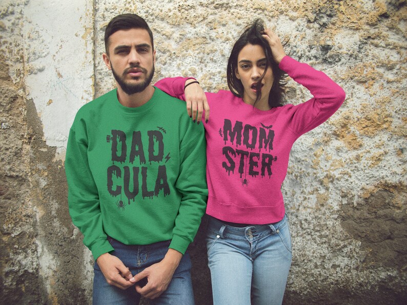 May include: A couple wearing matching green and pink sweatshirts with Halloween-themed text. The green sweatshirt says "Dad-Cula" and the pink sweatshirt says "Mom-ster".