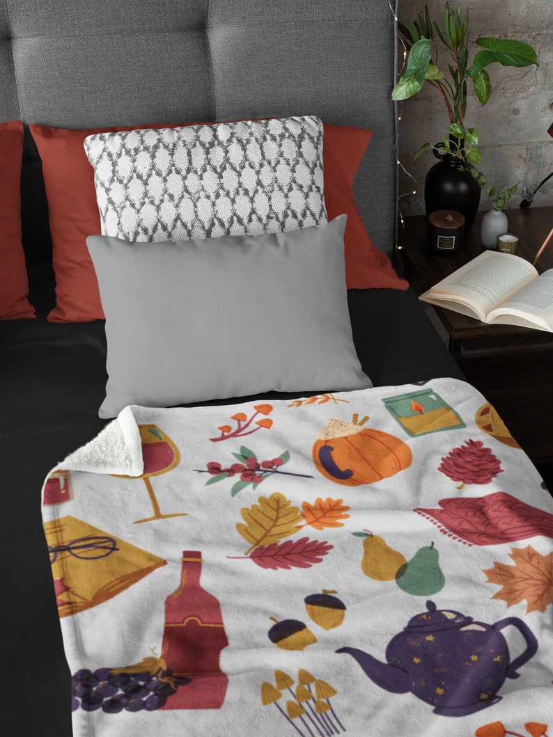Puede incluir: Una manta de forro polar blanca con un dise&ntilde;o de oto&ntilde;o. La manta presenta ilustraciones de calabazas, hojas, copas de vino, uvas, bellotas, setas, una tetera y un libro.
