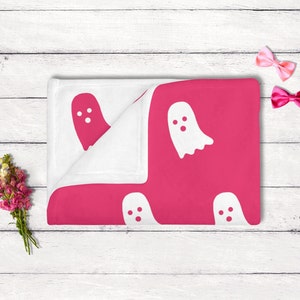 Puede incluir: Una manta de beb&eacute; rosa y blanca con un patr&oacute;n de fantasma. La manta est&aacute; doblada por la mitad y tiene un borde blanco.