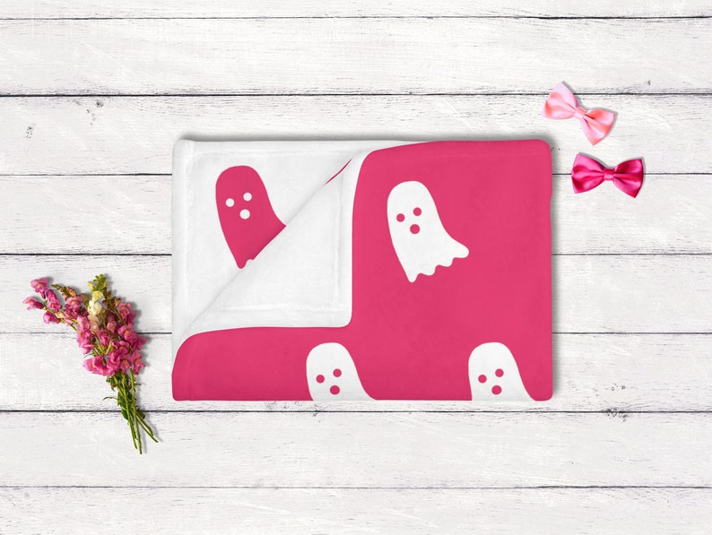 Puede incluir: Una manta de beb&eacute; rosa y blanca con un patr&oacute;n de fantasma. La manta est&aacute; doblada por la mitad y tiene un borde blanco.