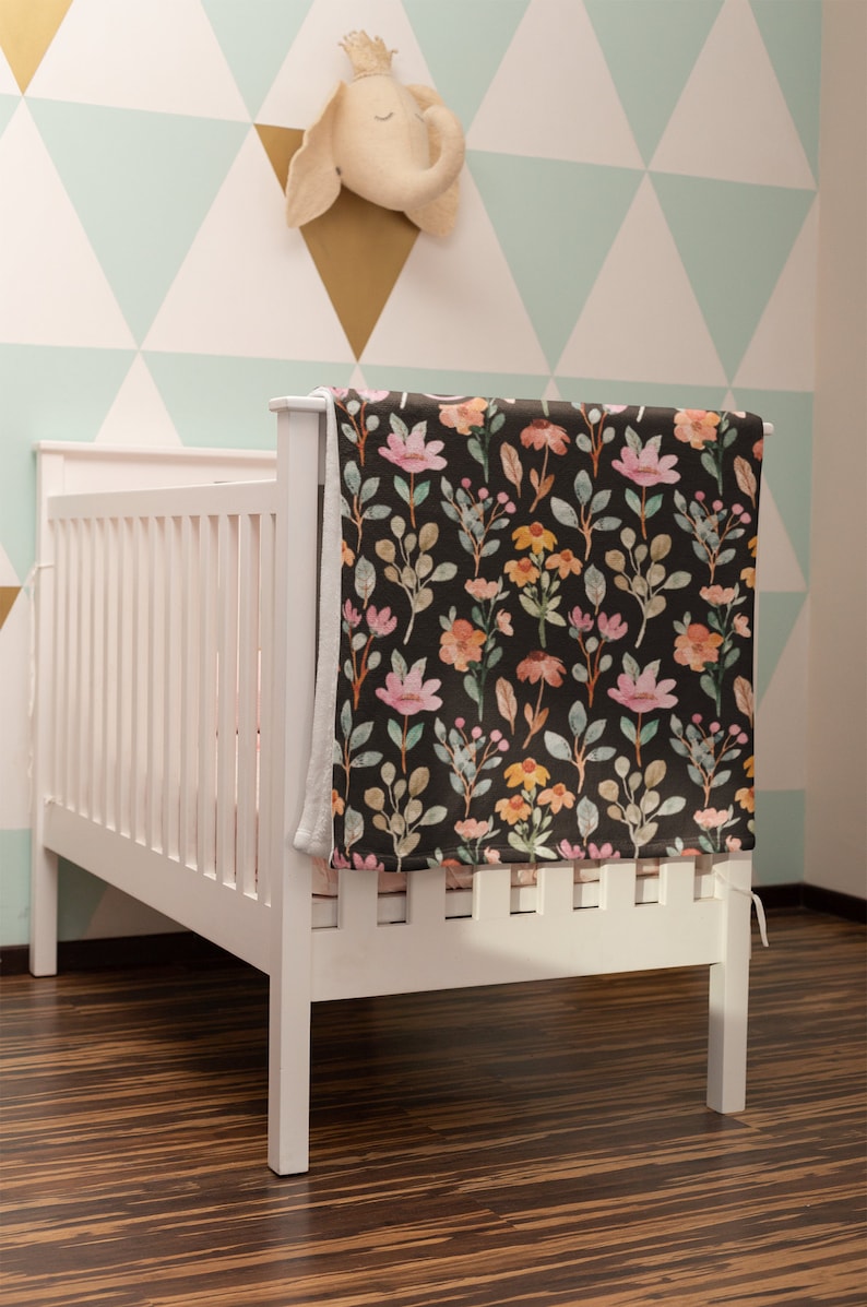 May include: A white crib with a floral patterned blanket draped over the side. The blanket features a dark background with pink, orange, and yellow flowers and green leaves. A plush elephant head decorates the wall.