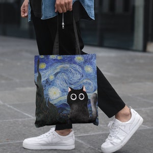 May include: A black tote bag with a black cat and a starry night painting design. The bag has black straps and is being held by a person.