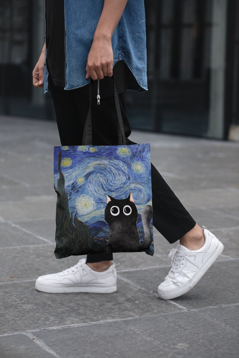 May include: A black tote bag with a black cat and a starry night painting design. The bag has black straps and is being held by a person.