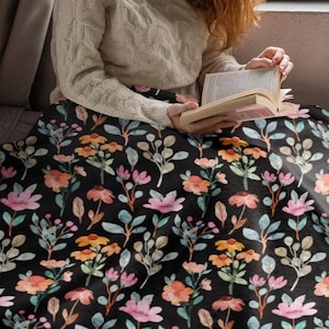May include: A black blanket with a floral pattern featuring pink, orange, and yellow flowers, along with green and blue leaves. The blanket is draped over a person, suggesting a cozy and comfortable item for home decor.