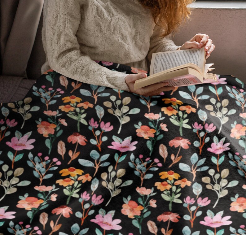 May include: A black blanket with a floral pattern featuring pink, orange, and yellow flowers, along with green and blue leaves. The blanket is draped over a person, suggesting a cozy and comfortable item for home decor.