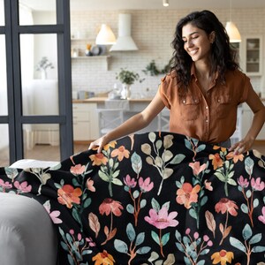 May include: A black throw blanket with a floral pattern featuring pink, orange, and yellow flowers, along with green and brown leaves. The blanket is being placed on a white sofa. The background shows a kitchen and a person.