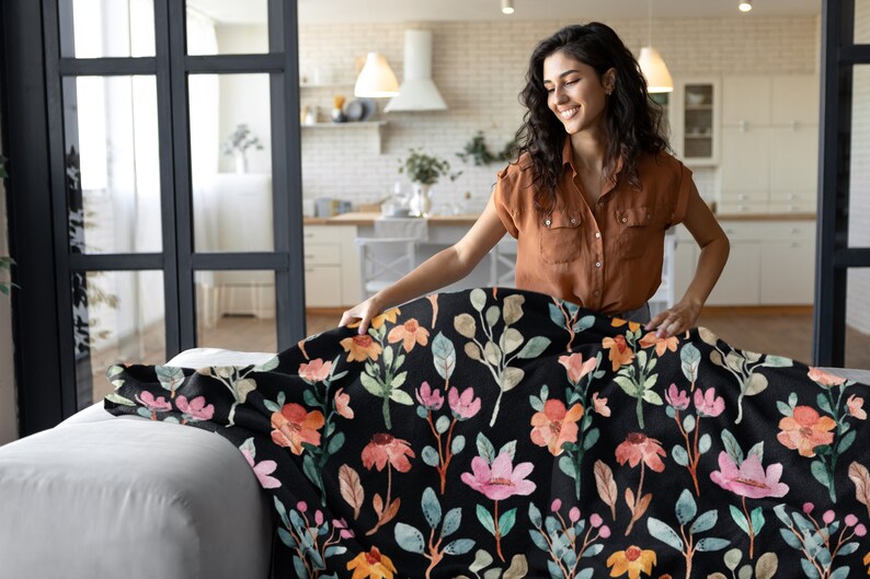 May include: A black throw blanket with a floral pattern featuring pink, orange, and yellow flowers, along with green and brown leaves. The blanket is being placed on a white sofa. The background shows a kitchen and a person.