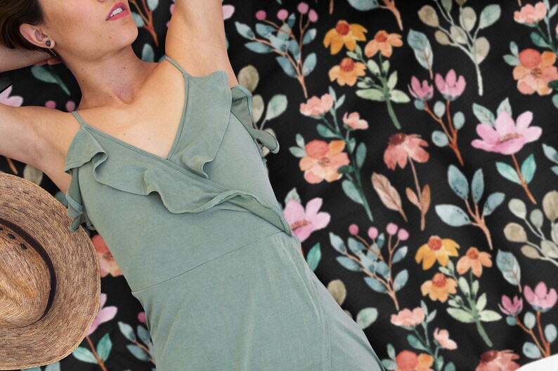 May include: A black floral patterned blanket with pink, orange, and yellow flowers and green leaves. A woman wearing a sage green dress and a straw hat is lying on the blanket.