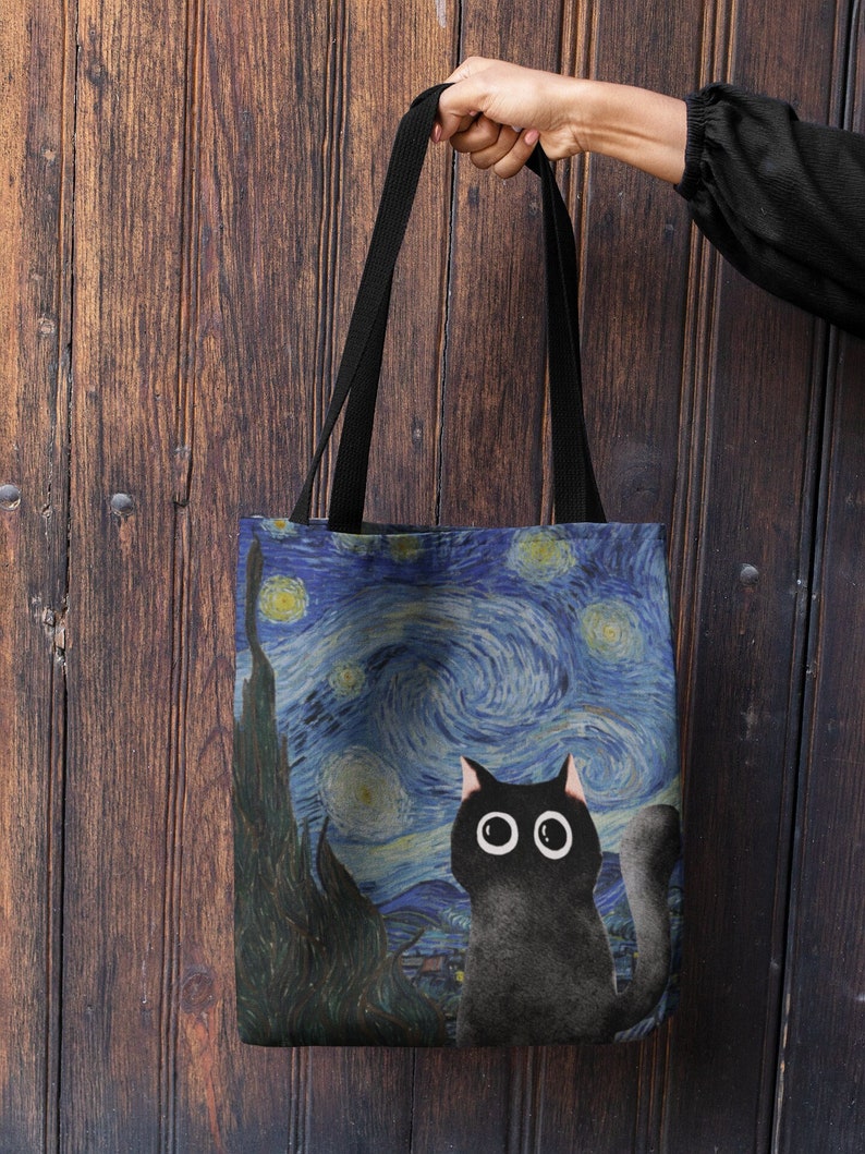 May include: A black tote bag with a black cat and a starry night painting. The cat has large, white eyes and is looking up. The painting is a recreation of Vincent van Gogh's "Starry Night."
