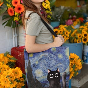 May include: A black tote bag with a print of Van Gogh's Starry Night and a black cat with large eyes. The bag has a black strap and is being held by a person.