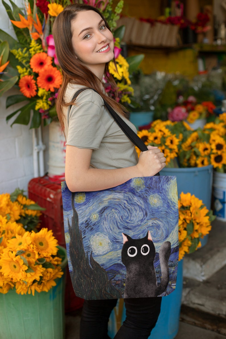 May include: A black tote bag with a print of Van Gogh's Starry Night and a black cat with large eyes. The bag has a black strap and is being held by a person.
