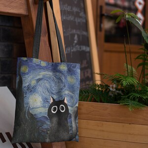 May include: A black tote bag with a blue and white starry night print featuring a black cat with large eyes. The bag has black straps.
