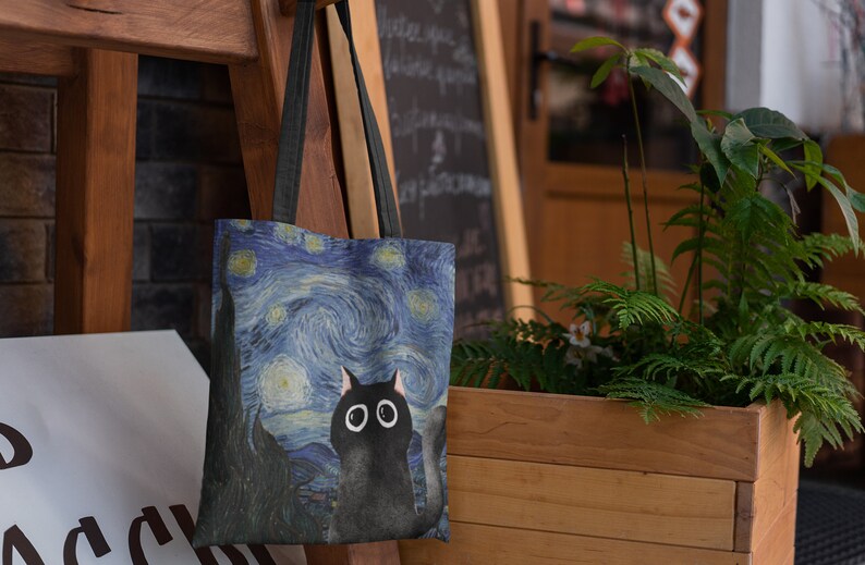 May include: A black tote bag with a blue and white starry night print featuring a black cat with large eyes. The bag has black straps.