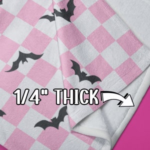 May include: Pink and white checkered fleece blanket with black bat silhouettes. The blanket is folded to show the 1/4 inch thickness.