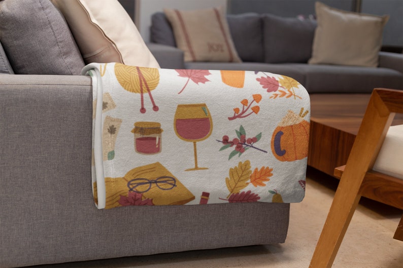 Puede incluir: Una manta blanca con un estampado colorido de tem&aacute;tica oto&ntilde;al. El estampado incluye hojas, calabazas, vasos de vino, tarros de mermelada, agujas de tejer y libros.