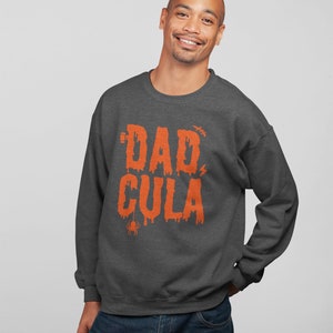 May include: A dark gray sweatshirt with a graphic print of the text "DAD-CULA" in orange. The text is dripping and has a spider and lightning bolt graphic.