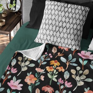 May include: A black blanket with a floral pattern featuring pink, orange, and yellow flowers, along with green and tan leaves. The blanket is on a bed with a white patterned pillow and black pillowcases. The blanket's underside is white.